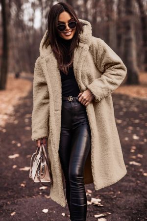 Femme élégante portant un manteau en fausse fourrure beige, lunettes de soleil, pantalon en cuir noir, tenant un sac à main, marchant dans un parc automnal.